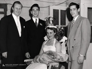A seated crowned woman holding a bouquet poses indoors with three men in suits.