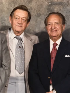 Two older men in formal suits pose side by side against a gray mottled studio backdrop.